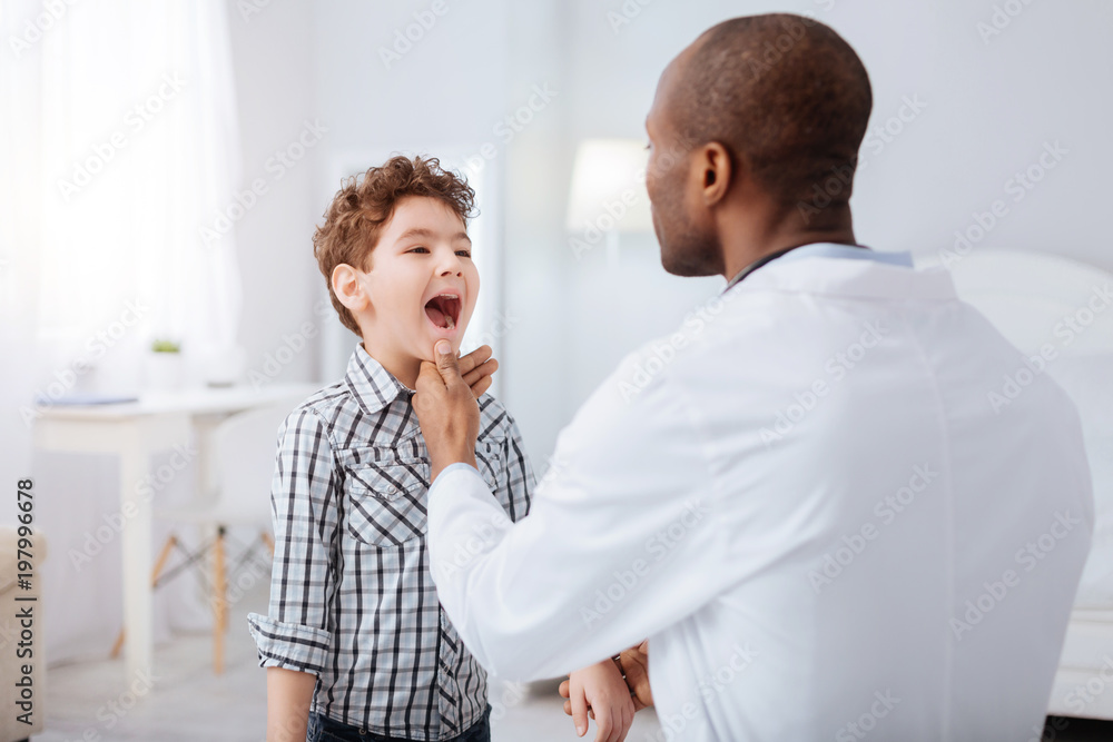 Strep throat. Pretty nice boy standing on blurred background while showing throat to doctor and waiting for instructions Strep throat. Pretty nice boy standing on blurred background while showing throat to doctor and waiting for instructions