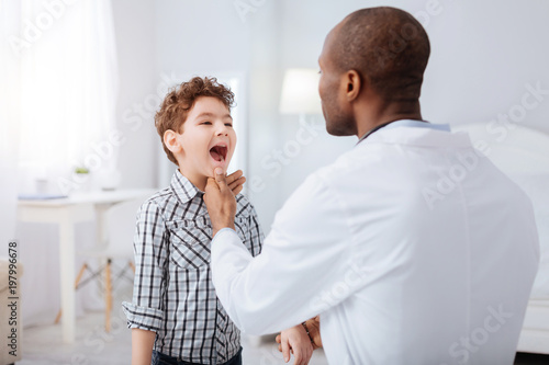 Strep throat. Pretty nice boy standing on blurred background while showing throat to doctor and waiting for instructions