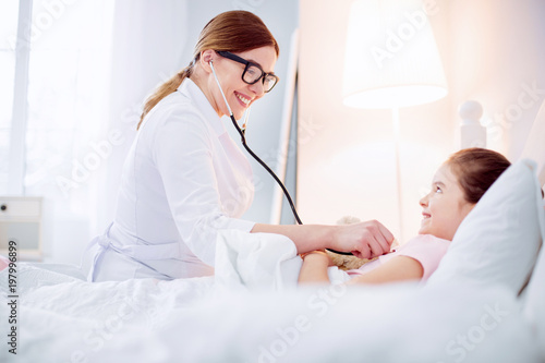 Good doctor. Optimistic vigorous female doctor hearing girl with stethoscope while smiling and looking at her