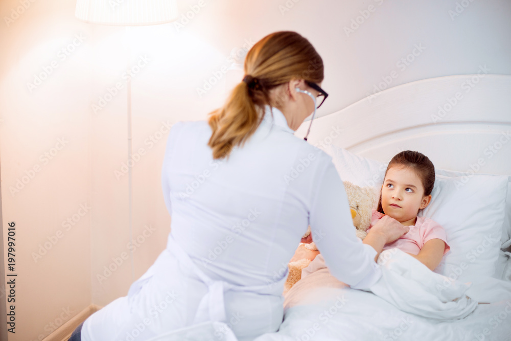 Just cold. Skillful female doctor hearing worried sad girl with stethoscope while she lying on bed and hugging plush bear Just cold. Skillful female doctor hearing worried sad girl with stethoscope while she lying on bed and hugging plush bear