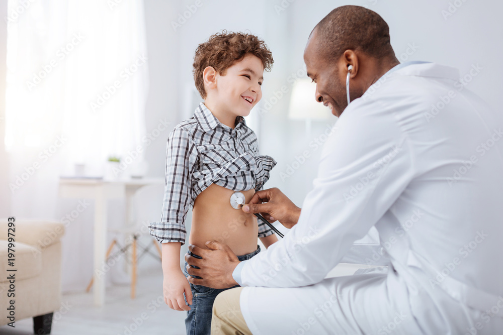 Medical tool. Cheerful merry boy laughing while male doctor using stethoscope and examining him Medical tool. Cheerful merry boy laughing while male doctor using stethoscope and examining him