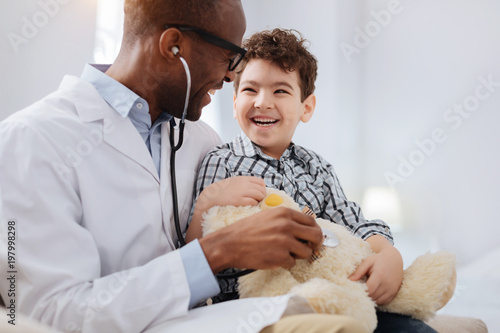 Immediate treatment. Enthusiastic vigorous male doctor sitting with boy while listening to plush bear and talking photo