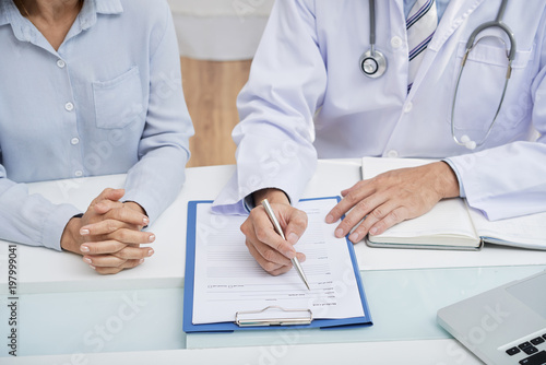 Close-up shot of male doctor wearing white coat filling in medical history while sitting at desk next to female patient