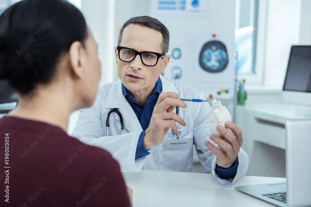 Explaining the problem. Professional therapeutist holdign a model of heart while having a check up Explaining the problem. Professional therapeutist holdign a model of heart while having a check up