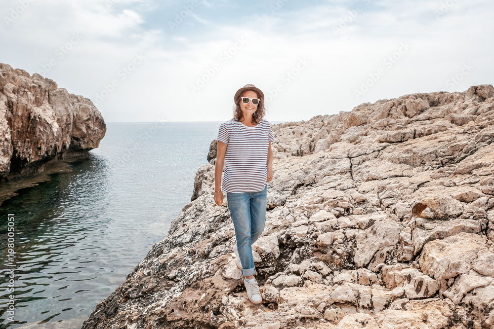 Young beautiful woman travel girl in hat and sunglasses walking along the rocky seashore, summer vacation and travel, sunny portrait