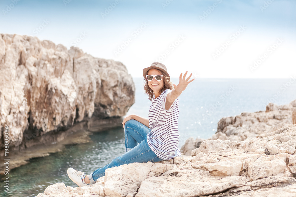 Young beautiful woman travel girl in hat and sunglasses sits on a rocky seashore, summer vacation and travel, sunny portrait