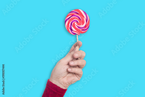 Left female hand holds colorful lollipop upright isolated on pastel light blue background - concept lollipop sweets sugar health tast food kids family fun