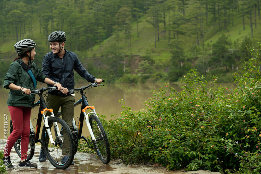 Naklejka premium Active Asian couple looking at each other while walking with their bicycles on rainy day