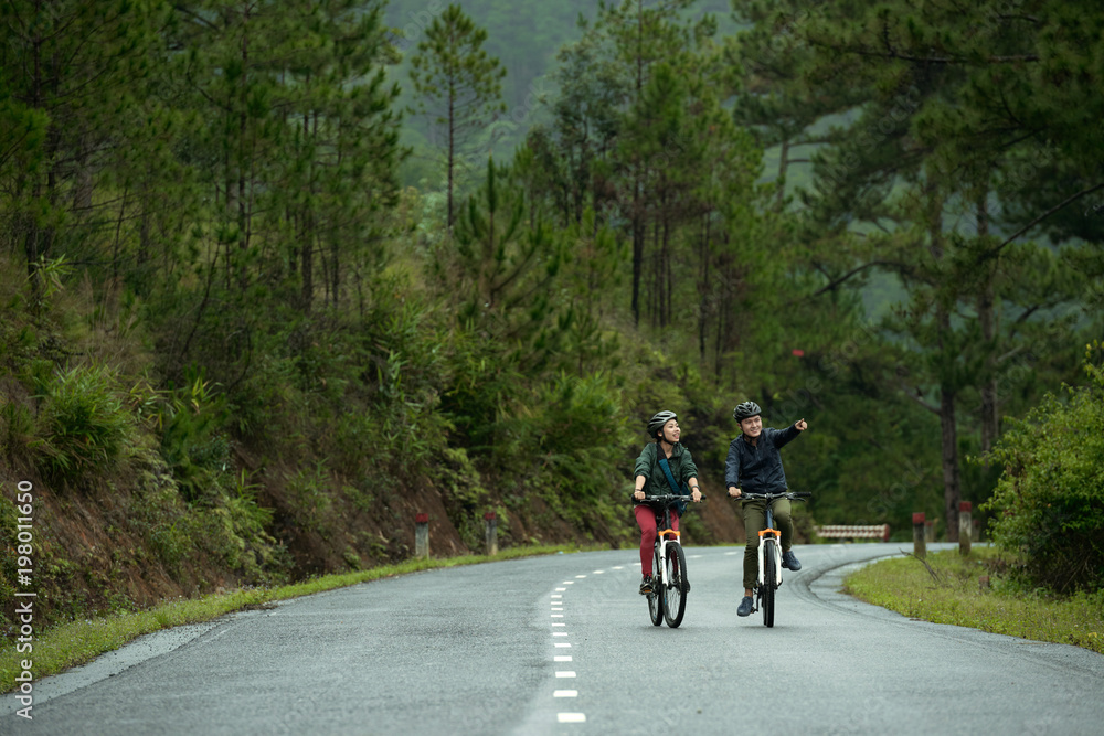 Asian couple cycling together down road, man pointing at something in the distance Asian couple cycling together down road, man pointing at something in the distance