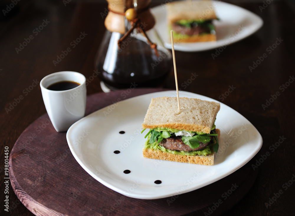 Burger with beef and greens in slices of unleavened bread. On white plate with drops of sauce. Coffee on background