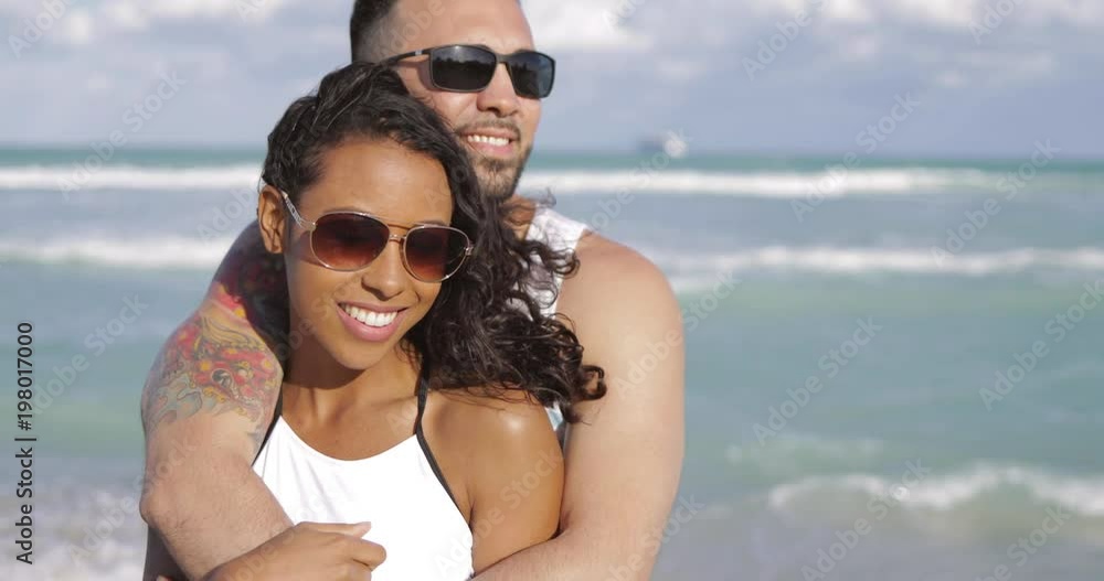 Happy handsome man embracing beautiful African-American girlfriend standing on background of ocean and looking away. Happy handsome man embracing beautiful African-American girlfriend standing on background of ocean and looking away.