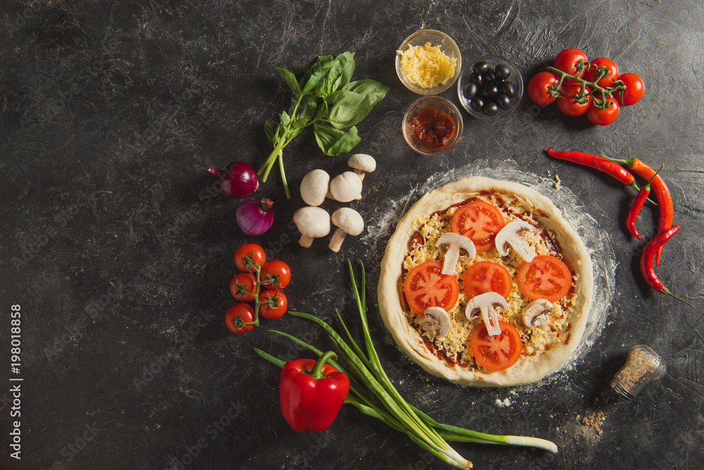 top view of raw italian pizza and fresh ingredients around on dark surface