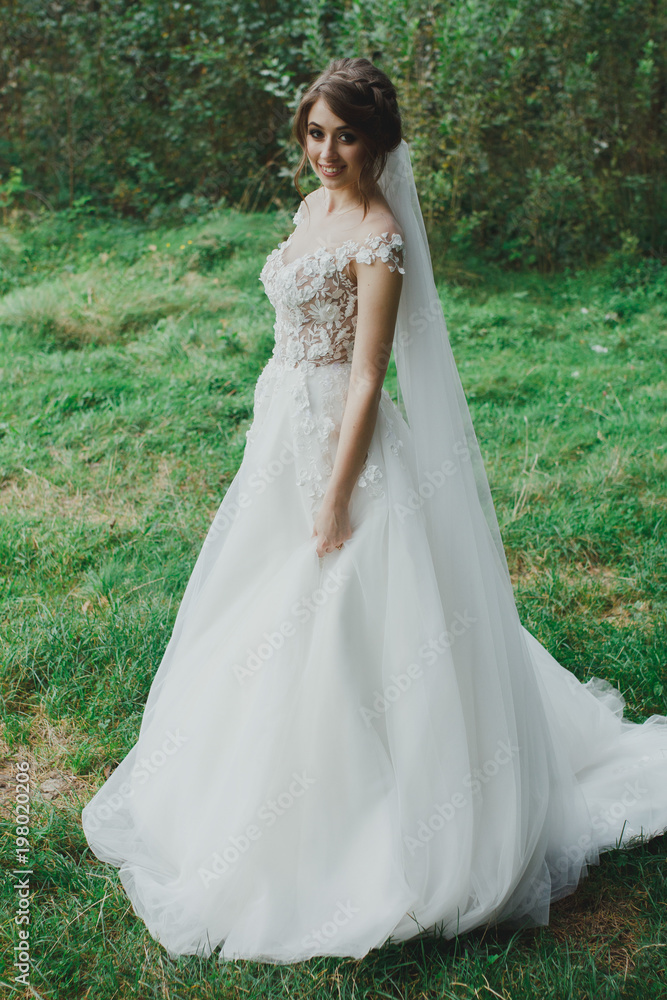 Beautiful bride with elegant simple hairdo is standing in the forest. Low open back white dress with flowers and tulle veil. Stylish hairstyle in outdoors romantic wedding photo. Bridal portrait.