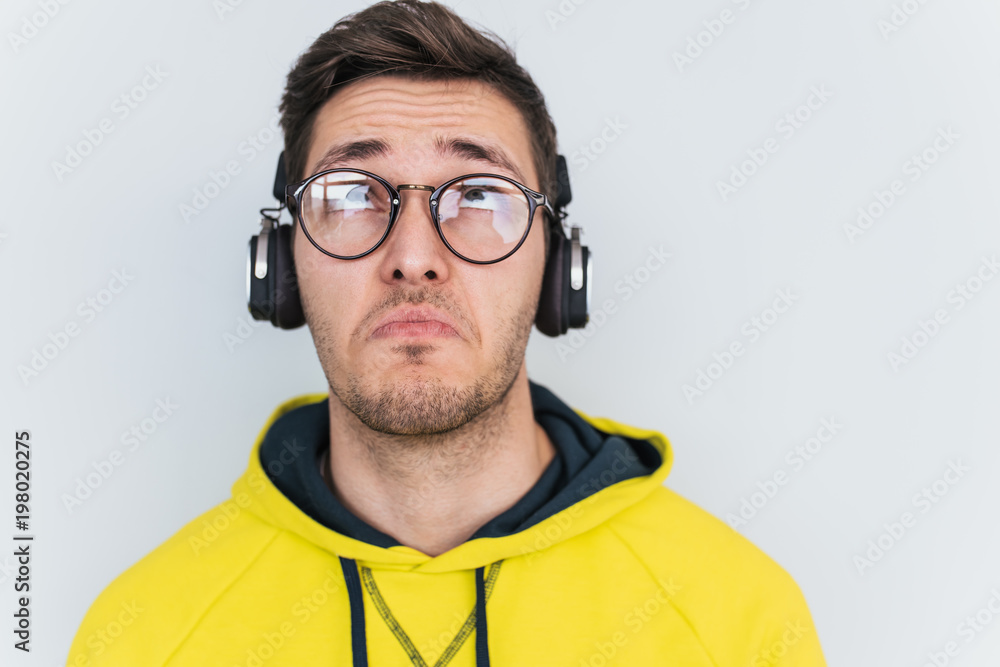 Studio horizontal portrait of frustrated man curves lower lips isolated over white studio background. Bearded male having sad expression wearing yellow hoodie and headphones. People, emotions concept