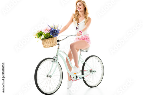 Young smiling girl riding bicycle with flowers in basket isolated on white