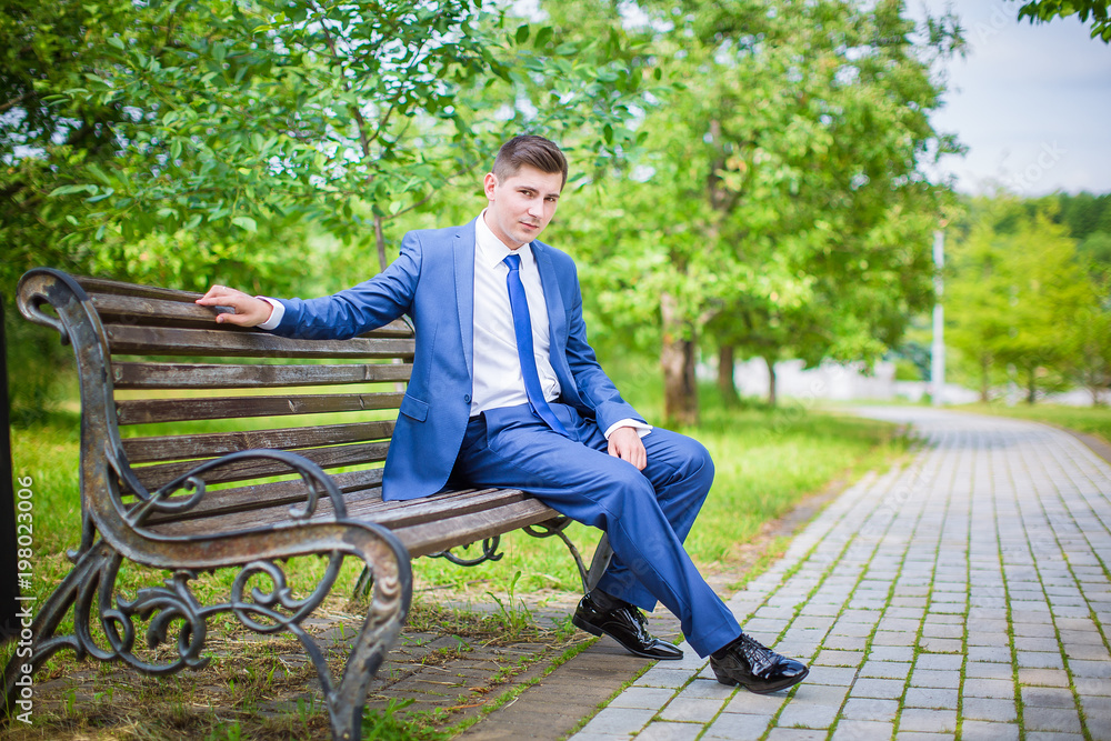 Obraz premium Stylish groom in blue suit, sitting on bench in park