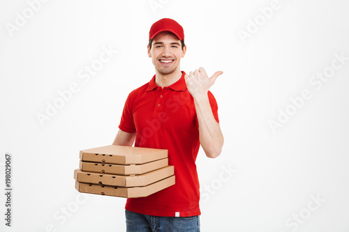 Photo of young man from delivery service 25y in red uniform carrying stack of pizza boxes and pointing finger aside on copyspace, isolated over white background photo