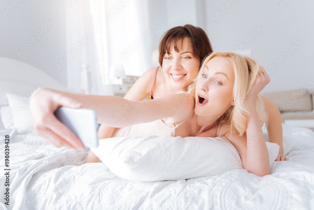 Pleasurable morning. Nice joyful female couple lying on the bed and taking selfies while being in a great mood Pleasurable morning. Nice joyful female couple lying on the bed and taking selfies while being in a great mood