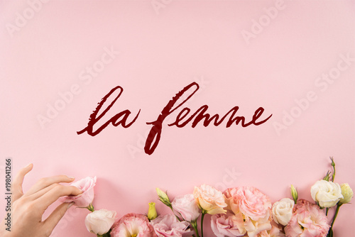 Top view of hand arranging beautiful tender flowers with LE FEMME sign isolated on pink background