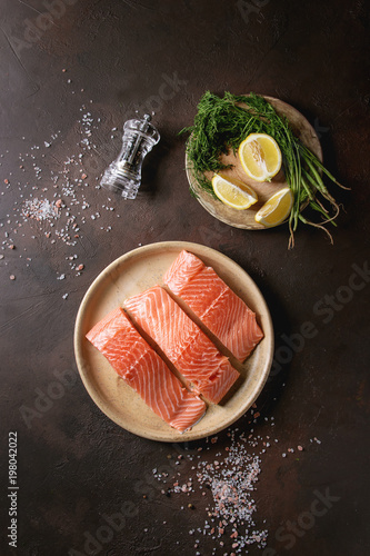 Sliced raw uncooked salmon fillet on ceramic plate with green dill, lemon, salt, pepper over dark brown texture background. Top view, space.