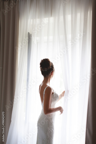 Beautiful bride in white wedding dress standing in her bedroom and looking in window. Beautiful young bride in white wedding dress indoors