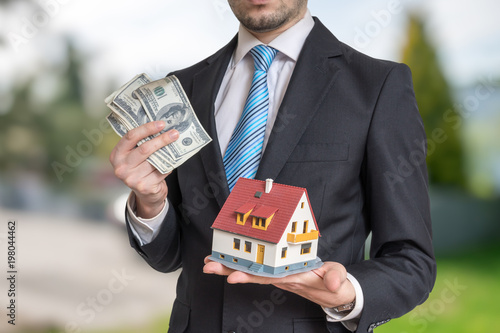 Real estate agent is holding small house and money in hands.