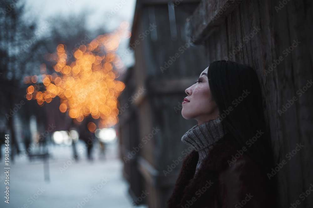 Portrait of young beautiful dreaming Asian girl woman in brown knitted sweater and boho leather coat walking outdoors in winter park or street with magic lights decorations and trees