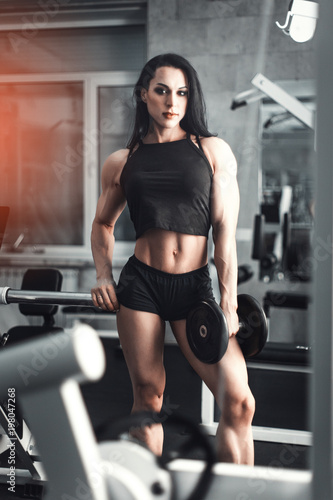 Full length portrait of a young attractive sexy beautiful fitness girl in black tank top and shorts standing in gym near training apparatus showing her press and looking at camera. Daria Manyilenkova