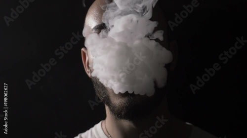 Bold young male musician vaper with beard vapes and blows on his mouth a huge cloud of vape smoke that covers his face in slow motion photo