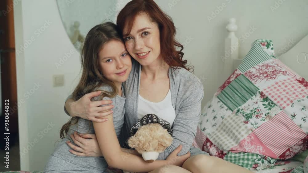 Portrait of adorable smiling girl embracing her happy mother and looking at camera together while sitting on bed in bright bedroom at home Portrait of adorable smiling girl embracing her happy mother and looking at camera together while sitting on bed in bright bedroom at home