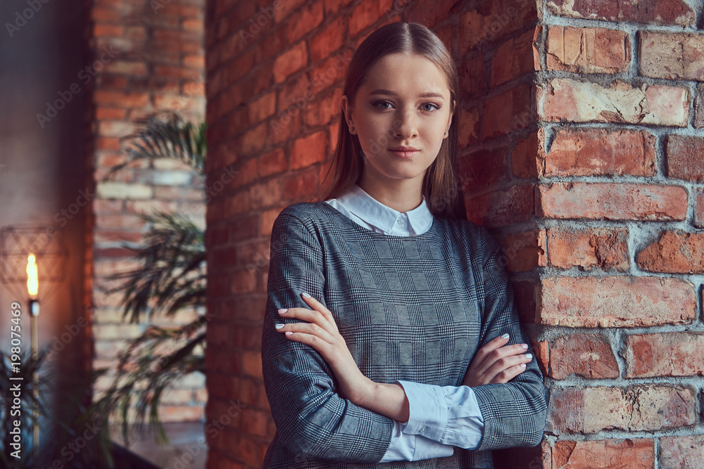 Portrait of a young slim sensual girl in a gray dress leaning against a brick wall with a crossed arms.