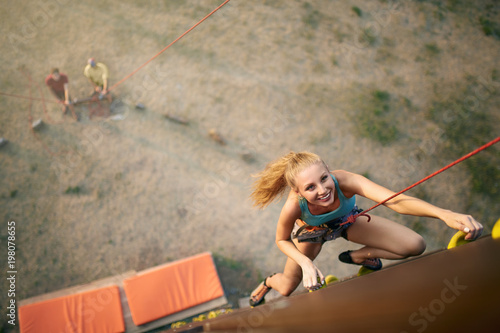 Beautiful young strong woman climbing on rock artificial wall in summer, top view. Climber insured on belaying harness by friends reaches top of the cliff outdoors.