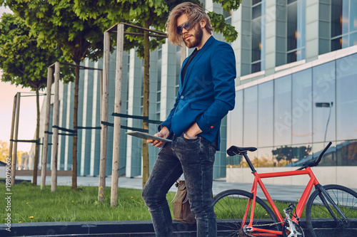 A handsome fashionable bearded male with long hair dressed in stylish clothes, working with a tablet computer after bicycle riding.