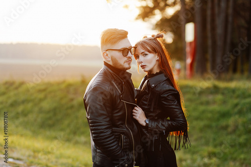 slim stylish girl with long red hair hugging a brutal guy with a beard and wearing sunglasses at sunset