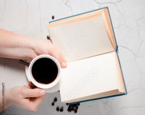 Fried coffee beans and in the hands of the girl a cup of hot delicious coffee and an open book on a gray background. Place for text. Concept of coffee beans