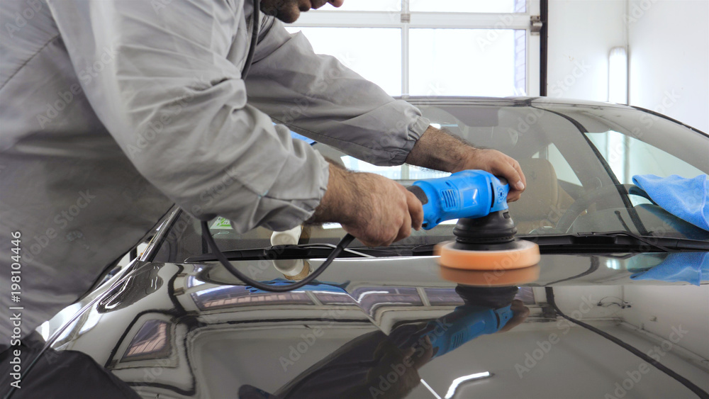 A man in a special suit polishes a gray car body, a tool for polishing cars, into a workshop. A man in a special suit polishes a gray car body, a tool for polishing cars, into a workshop.