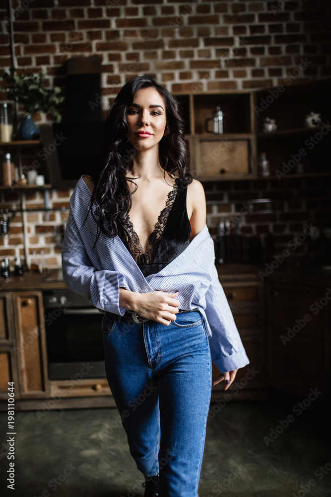 Fototapeta premium Beautiful brunette in the kitchen, modern housewife, fashion, wooden brown furniture, dressed in blue jeans and a blue shirt