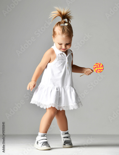 Young pretty toddler girl kid with big sweet lollypop candy in white dress on grey
