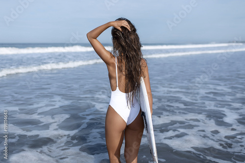 Fototapeta Naklejka Na Ścianę i Meble -  Surfer girl in a white swimsuit goes to the blue ocean, with a white surfboard