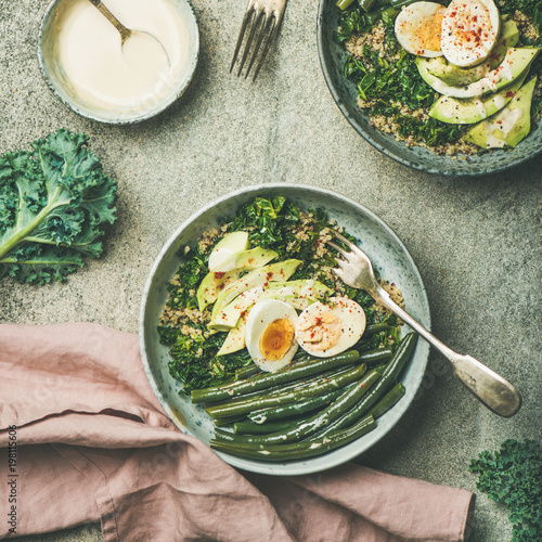 Healthy vegetarian breakfast bowls flat-lay. Quinoa, kale, green beans, avocado, egg and tahini dressing bowls over concrete background, top view, square crop. Energy boosting, diet food concept