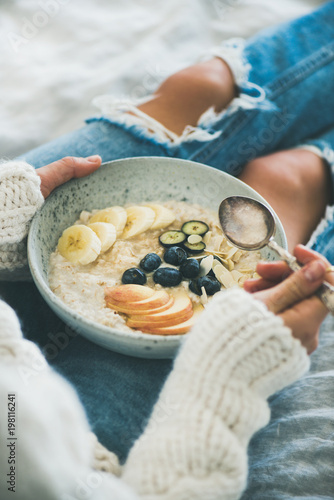 Healthy winter breakfast in bed. Woman in woolen sweater and shabby jeans eating vegan almond milk oatmeal porridge with berries, fruit and almonds, close-up. Clean eating, vegetarian food concept
