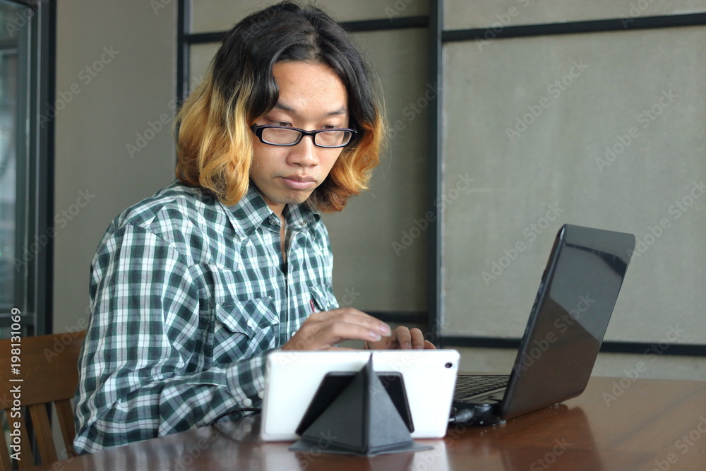 Relaxed young Asian hipster man working against laptop and tablet in his office.