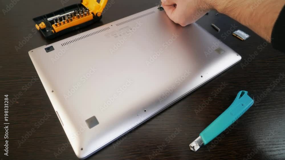 The master fastens the back cover of the laptop with screws. Close-up hands tighten the screw using a screwdriver. The master fastens the back cover of the laptop with screws. Close-up hands tighten the screw using a screwdriver.
