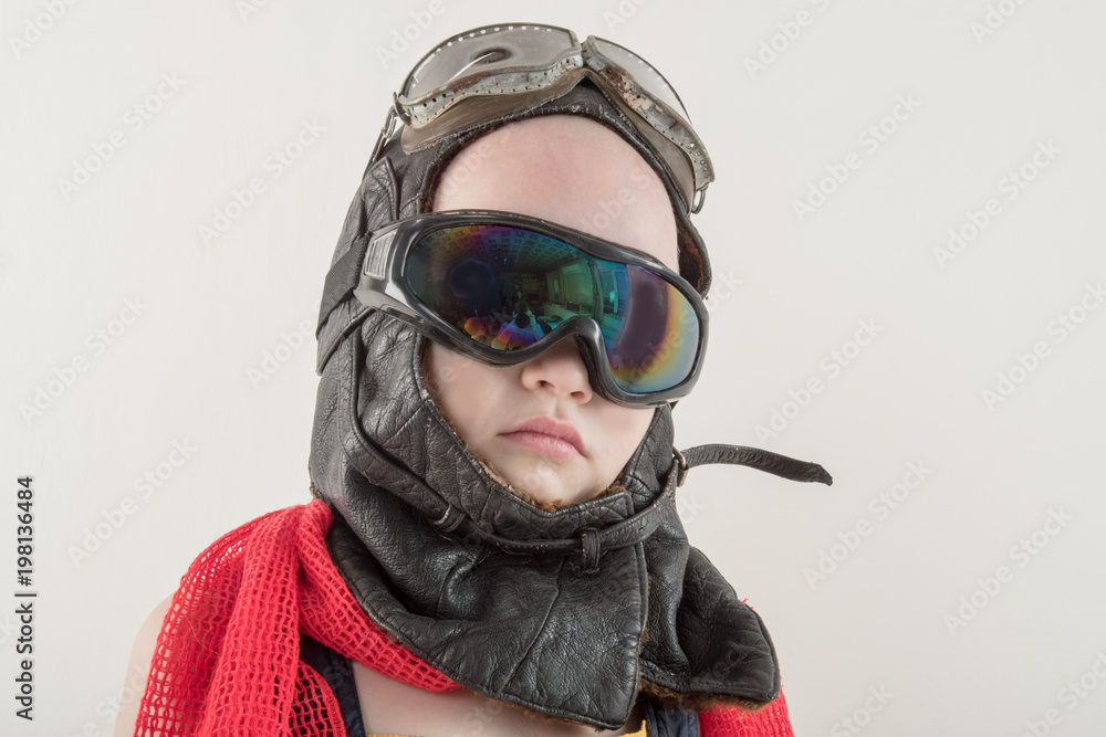 Funny boy in yellow overalls and hat, in a red scarf and pilot's goggles, sits on vintage suitcase with an airplane model and dreams of adventures. The concept of travel romance and discovery Funny boy in yellow overalls and hat, in a red scarf and pilot's goggles, sits on vintage suitcase with an airplane model and dreams of adventures. The concept of travel romance and discovery