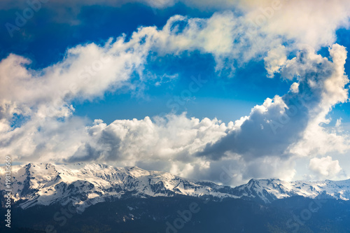 A mountain range with thawing snow and ice in the spring, against the background of forest slopes under a blue sky in the clouds. Ski Resort at Caucasus Mountains, Rosa Peak, Sochi, Russia.