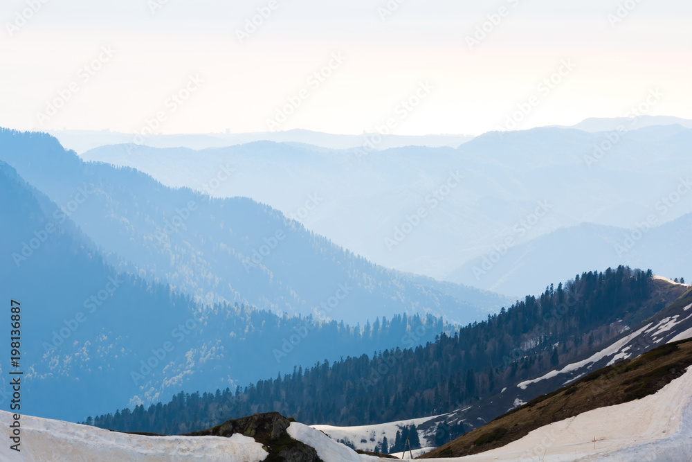 A mountain range with thawing snow and ice in the spring, against the background of forest slopes under a blue sky in the clouds. Ski Resort at Caucasus Mountains, Rosa Peak, Sochi, Russia.