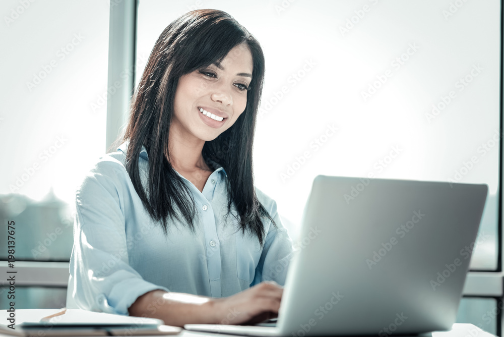 Best work. Pretty occupied responsible employee sitting by the table smiling and using the laptop. Best work. Pretty occupied responsible employee sitting by the table smiling and using the laptop.