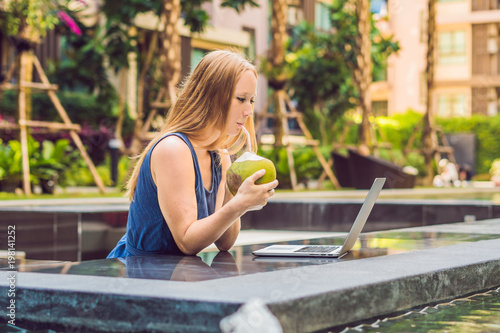 Young female freelancer sitting near the pool with her laptop in the hotel browsing in her smartphone. Busy at holidays. Distant work concept. Copy space for your text photo