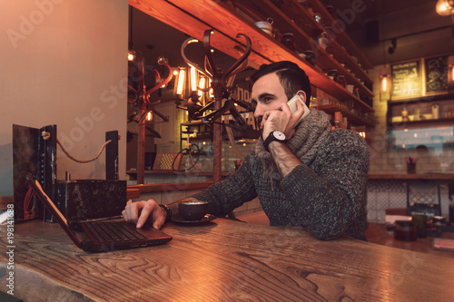 A young man with a beard sits at a table in a cafe and works behind a small laptop with a cup of delicious coffee