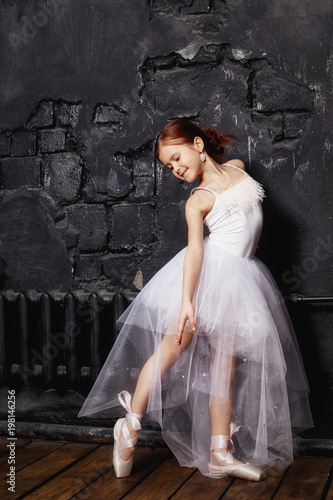 Girl in a white ball gown and shoes, beautiful red hair. Young theater actress. Little prima ballet. Young ballerina girl is preparing for a ballet performance